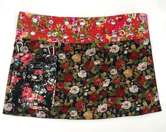 May include: A floral print apron with a red top band and a navy blue skirt. The apron features a pocket with a black floral print and silver grommets. The apron is designed for use in the kitchen or for crafting.