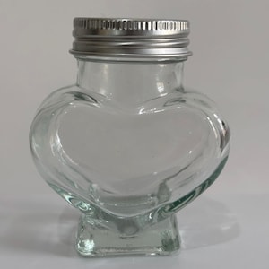 May include: A clear glass heart-shaped jar with a silver metal lid. The jar has a square base and is empty. The jar is suitable for storing small items or as a decorative piece.