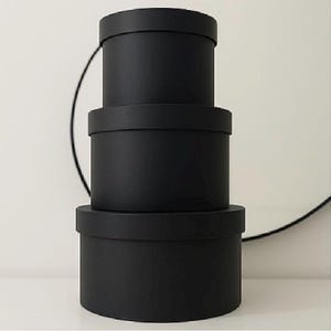 May include: Three black, round gift boxes stacked on top of each other. The boxes are of different sizes, with the largest at the base and the smallest at the top. They have matching lids and a matte finish. The boxes are a uniform black colour.