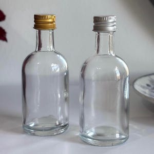 May include: Two clear glass bottles with rounded bodies and narrow necks. One has a gold cap, the other a silver cap. The bottles are empty, suitable for small liquid portions. Each bottle is approximately 15 cm tall.