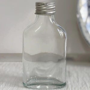 May include: A clear, rectangular glass bottle with a silver metal screw-on cap. The bottle has rounded corners and a flat base. The background is a soft white, highlighting the bottle's transparency. The bottle is empty.