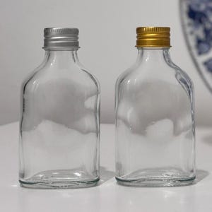 May include: Two clear glass bottles with silver and gold metal screw-on caps. The bottles have a flat, rectangular shape with rounded corners. The caps are ribbed and the bottles are empty.