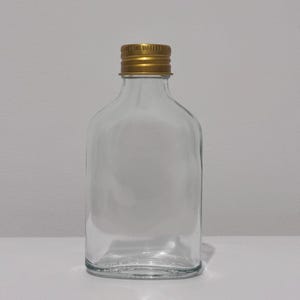 May include: A clear glass flask with a gold-coloured metal screw-on cap. The flask has a flat, rectangular body with rounded shoulders and a narrow neck. The bottle is empty and sits on a white surface.