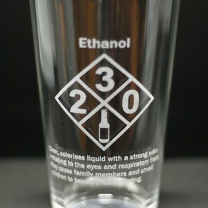 May include: A clear glass shot glass with the word "Ethanol" and a diamond-shaped warning symbol with the numbers "2", "3", and "0" etched on the front. The symbol also includes a small image of a bottle.