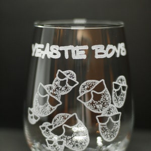 May include: A clear glass wine glass with the words "Yeastie Boys" etched on the side. The glass features a design of several cartoon-style characters wearing sunglasses.