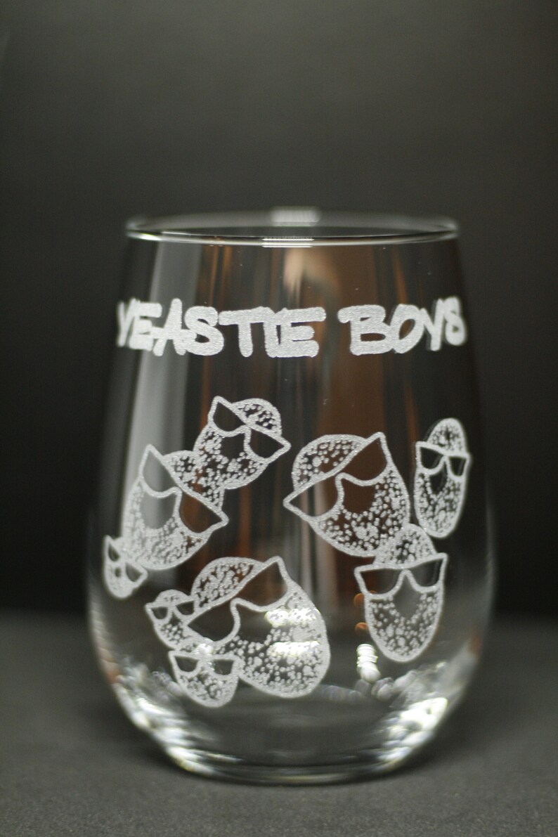 May include: A clear glass wine glass with the words "Yeastie Boys" etched on the side. The glass features a design of several cartoon-style characters wearing sunglasses.