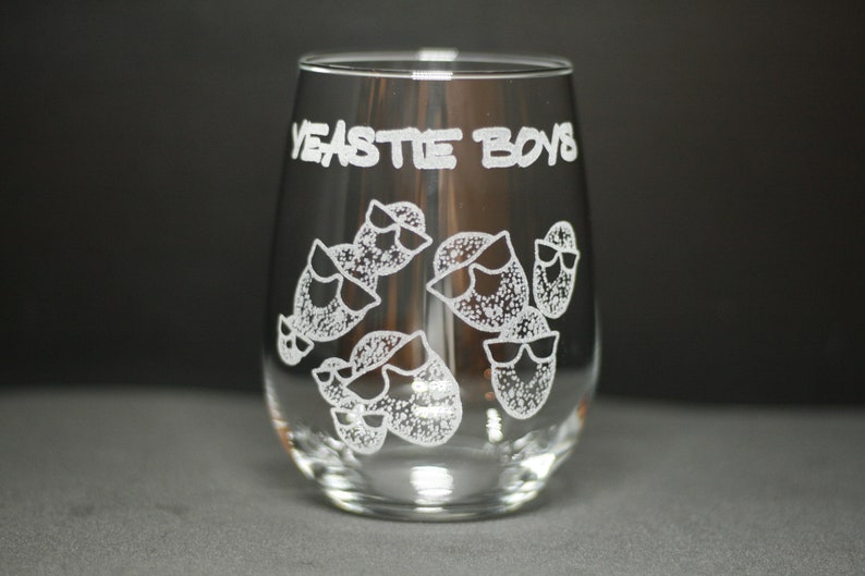May include: A clear glass wine glass with the text "YEASTIE BOYS" etched on the front. The glass features a design of multiple cartoon faces wearing sunglasses.