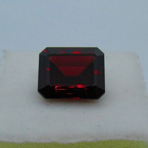 May include: A deep red gemstone with a rectangular emerald cut. The gemstone is set on a white surface.