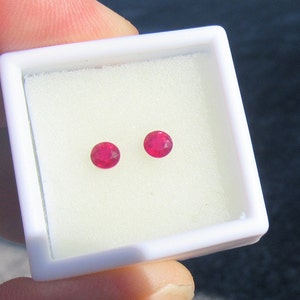 Puede incluir: Dos gemas redondas, facetadas, de color rojo brillante, se exhiben en una caja cuadrada blanca con un inserto de espuma blanca. Las gemas son de tamaño y forma similares, y probablemente sean rubíes.
