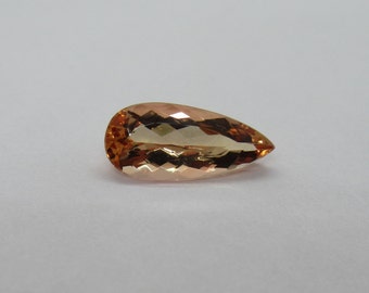 May include: A pear-shaped gemstone with a light peach and orange color. The gemstone has a faceted cut and is set against a white background.