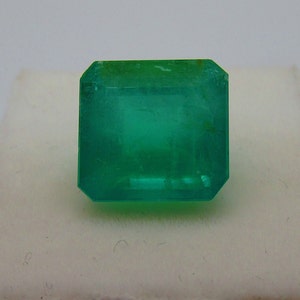 May include: A vibrant green emerald gemstone, cut into a square shape with bevelled edges. The emerald is set on a white background.