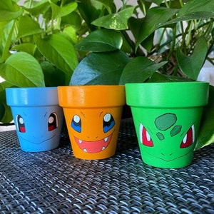 Peut inclure: Trois petits pots de fleurs en terre cuite peints avec des visages de dessins animés des personnages Pokémon Bulbizarre, Salamèche et Carapuce. Les pots sont disposés en rangée sur une surface tissée noire et grise.