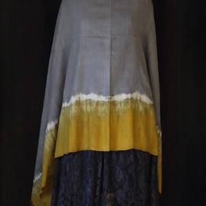 May include: A gray shawl with a white and yellow ombre design is draped over a dress form. The shawl has a fringed edge and a dark blue lace skirt is visible below. The background is black.