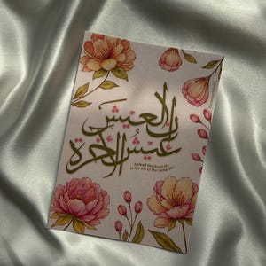 May include: A rectangular card featuring a light pink background with watercolour-style illustrations of pink and orange flowers. The card displays dark green Arabic calligraphy and the English text: Indeed the (true) life is the life of the hereafter.