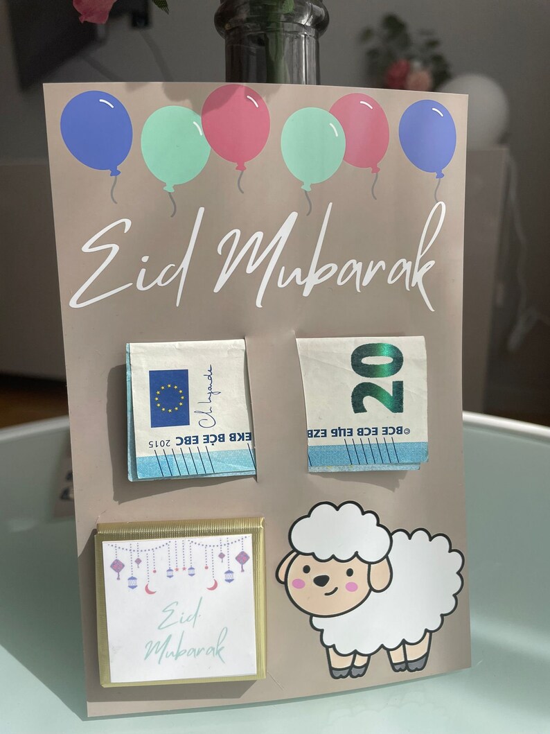 May include: A light brown card with the text "Eid Mubarak" in white cursive writing. There are five colorful balloons at the top of the card. The card has two 20 euro bills, a chocolate bar with the text "Eid Mubarak" and a cartoon white sheep.