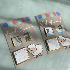May include: Two cards with a light brown background and colorful balloons at the top. The card on the left has the text "Hayırlı Bayramlar" and a picture of a white sheep. The card on the right has the text "Eid Mubarak" and a picture of a white sheep. Both cards have a small square with a gold frame and text inside. The card on the left has the text "Hayırlı Bayramlar" and the card on the right has the text "Eid Mubarak".