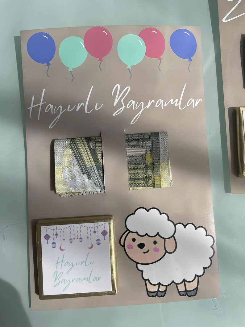 May include: A light brown card with the text "Hayirli Bayramlar" in white. The card is decorated with blue, pink, and green balloons. There are two folded bills of money on the card. There is a small gold frame with the text "Hayirli Bayramlar" in purple and a white cartoon sheep.