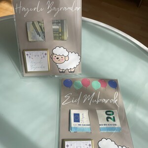 May include: Two cards with a light brown background and a white sheep illustration. The top card has the text "Hayırlı Bayramlar" and the bottom card has the text "Eid Mubarak". Both cards have colorful balloons and a small square with the text "Hayırlı Bayramlar" and "Eid Mubarak" respectively.