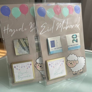 May include: Two gift envelopes with a brown background and colorful balloons. The envelopes have a gold border and a small square window with a cash gift inside. The envelopes have the text "Hazırlı Bayrandar" and "Eid Mubarak" printed on them.