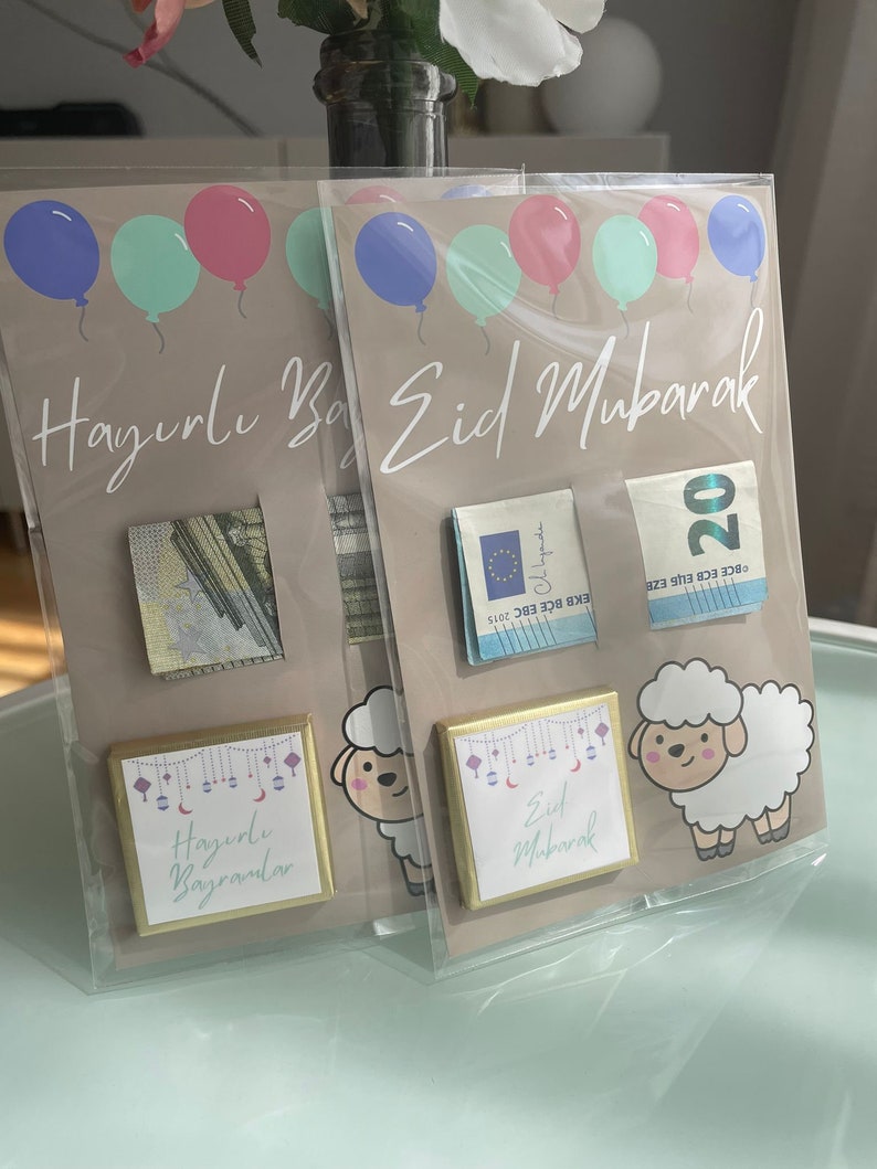 May include: Two gift envelopes with a brown background and colorful balloons. The envelopes have a gold border and a small square window with a cash gift inside. The envelopes have the text "Hazırlı Bayrandar" and "Eid Mubarak" printed on them.
