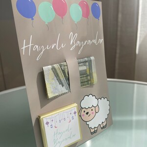 May include: A light brown card with the text "Hayırlı Bayramlar" in white. The card has five balloons in blue, pink, and green. There are two gold envelopes with a pattern of stars and a white sheep with pink cheeks. The card also has a small chocolate bar with the text "Hayırlı Bayramlar" in white.