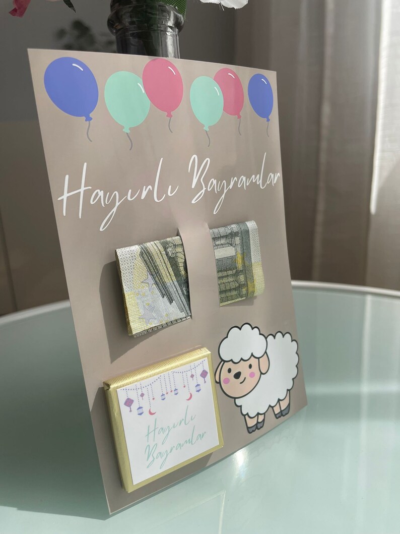 May include: A light brown card with the text "Hayırlı Bayramlar" in white. The card has five balloons in blue, pink, and green. There are two gold envelopes with a pattern of stars and a white sheep with pink cheeks. The card also has a small chocolate bar with the text "Hayırlı Bayramlar" in white.