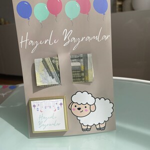 May include: A light brown card with the text "Hayırlı Bayramlar" written in white. The card has five balloons in the top left corner, two pink, two light blue, and one dark blue. There are two small squares with money inside, and a small square with a gold border and the text "Hayırlı Bayramlar" inside. There is a white cartoon sheep on the right side of the card.
