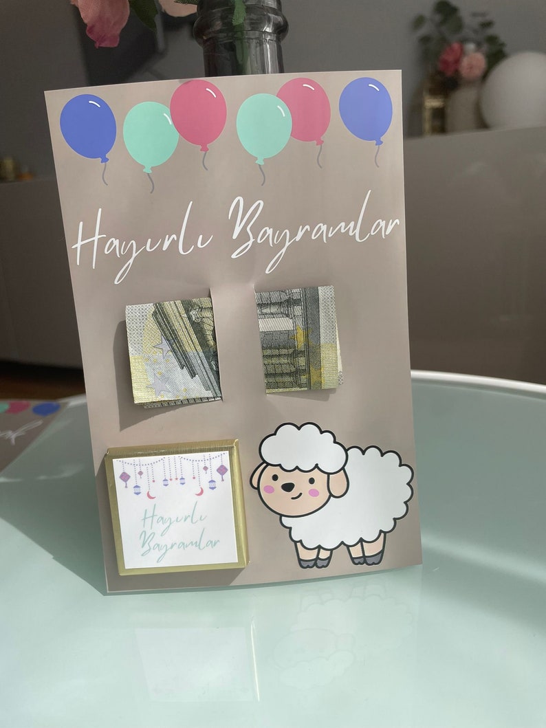 May include: A light brown card with the text "Hayırlı Bayramlar" written in white. The card has five balloons in the top left corner, two pink, two light blue, and one dark blue. There are two small squares with money inside, and a small square with a gold border and the text "Hayırlı Bayramlar" inside. There is a white cartoon sheep on the right side of the card.