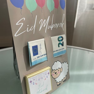 May include: A brown card with the text "Eid Mubarak" in white cursive writing. There are five balloons in the top row, two blue, two green, and one pink. There are three items attached to the card: a chocolate bar with a gold wrapper and the text "Eid Mubarak" in white cursive writing, a twenty euro bill, and a small piece of paper with the text "Eid Mubarak" in blue cursive writing.