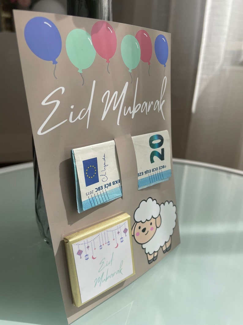 May include: A brown card with the text "Eid Mubarak" in white cursive writing. There are five balloons in the top row, two blue, two green, and one pink. There are three items attached to the card: a chocolate bar with a gold wrapper and the text "Eid Mubarak" in white cursive writing, a twenty euro bill, and a small piece of paper with the text "Eid Mubarak" in blue cursive writing.