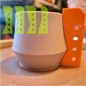 May include: A light-colored ceramic cup with a rounded base and a wider top. An orange trimming tool is positioned next to the cup. Green stencils are overlaid on the cup, suggesting a design process. The cup is on a light-colored surface.