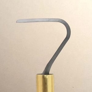 Peut inclure: Un outil de sculpture en métal doté d'une lame incurvée à bout plat en acier gris foncé. La lame est fixée à une virole cylindrique en métal doré, reliée à un manche noir lisse qui s'affine progressivement vers la base de l'outil.