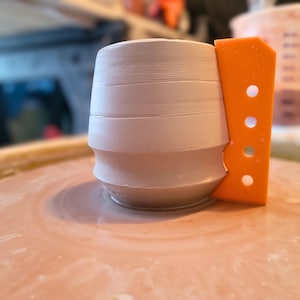 May include: A light grey, cylindrical clay vessel centred on a pottery wheel, featuring horizontal ridges along its exterior. An orange plastic rib tool with four circular cutouts is positioned vertically against the side of the clay form.