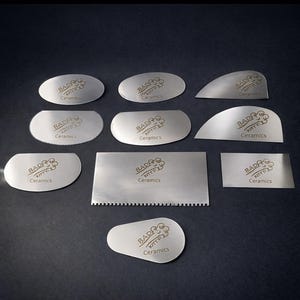 May include: A collection of stainless steel ceramic tools in various shapes, including oval, curved, and rectangular designs. Each tool is engraved with the text "BAD KITTY Ceramics". The tools are arranged on a dark surface.