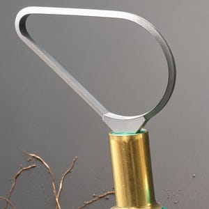 May include: A pottery trimming tool featuring a silver-coloured metal loop blade shaped like a teardrop, attached to a gold-toned cylindrical metal ferrule and a green handle. The tool is designed for shaping and refining clay surfaces.