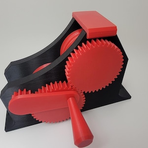 Pode incluir: Um mecanismo de engrenagem impresso em 3D preto e vermelho com uma alça vermelha. As engrenagens estão interligadas e giram quando a alça é girada.