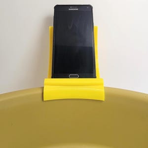 May include: A black Samsung smartphone is held in a bright yellow phone holder. The phone holder is curved and sits on a yellow surface. The phone's screen is black.
