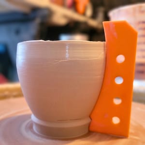 May include: A light beige ceramic cup with a smooth, rounded body and a flared base. Attached to the side is a bright orange, flat, vertical handle featuring four evenly spaced circular cutouts, creating a distinct contrast in color and form.