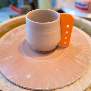 May include: A light-colored ceramic mug with an orange trimming tool attached, sitting on a pottery wheel. The mug is cylindrical with a rounded base. The orange tool has several holes. The pottery wheel is a light pink color.