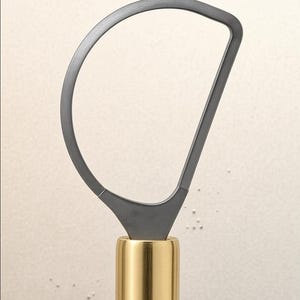 May include: A pottery trimming tool featuring a D-shaped dark gray metal loop blade securely attached to a cylindrical, polished gold-toned metal handle. The tool has a smooth, clean design with a distinct contrast between the matte gray loop and the shiny gold handle, showing a functional construction for clay shaping and trimming tasks.
