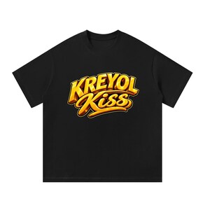 Puede incluir: Una camiseta negra de manga corta con un estampado gráfico en el pecho. El diseño muestra el texto KREYOL KISS en una fuente dorada metálica, audaz y tridimensional, con un contorno oscuro sobre el tejido negro de la prenda.