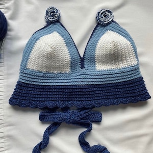 May include: A blue, white, and light blue crocheted crop top with a scalloped edge and a tie closure. The top has a floral design on the straps.