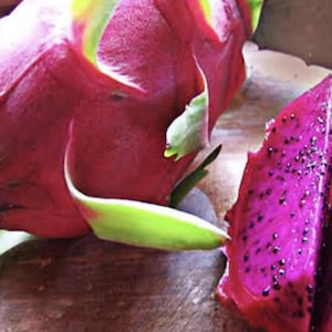 May include: A whole dragon fruit with vibrant pink skin and green-tipped scales positioned next to a sliced piece of dragon fruit, revealing a deep magenta flesh speckled with numerous small, edible black seeds throughout the interior.