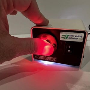 Könnte beinhalten: Ein kleines, rechteckiges Gerät mit rotem Licht, das von einem Drehknopf ausgeht. Das Gerät hat eine schwarze Front mit einem Netzschalter und einem Etikett mit der Aufschrift "Solarc Lighting Technology". Das Gerät ist weiß mit rosa Verkleidung.