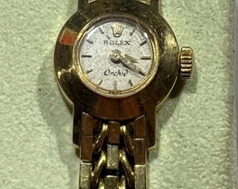 May include: A gold-toned Rolex Orchid wristwatch with a textured dial and a matching gold bracelet. The watch face displays the Rolex logo and the word "Orchid". The watch is set against a light green background.