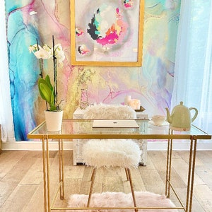 May include: A gold metal desk with a glass top and a white fluffy chair. The desk is in front of a wall with a colorful abstract painting and a white orchid plant.