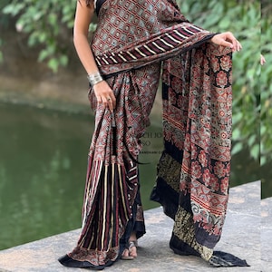 Pu&ograve; includere: Un lungo sari drappeggiato con una base scura e un motivo geometrico a rombi ripetuto in rosso, bianco e nero. Il tessuto presenta sezioni con strisce verticali e motivi floreali, rifinito con un bordo nero solido lungo i margini.