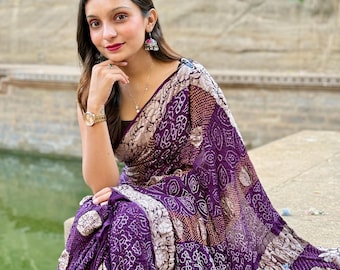 Può includere: Una donna indossa un sari a fantasia viola e bianco, seduta vicino all'acqua. Il sari presenta intricati disegni floreali e geometrici nei toni del viola, bianco e marrone. La donna indossa gioielli, tra cui orecchini e un orologio.