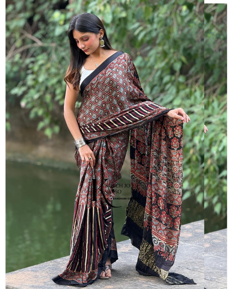 Pu&ograve; includere: Un lungo sari drappeggiato con una base scura e un motivo geometrico a rombi ripetuto in rosso, bianco e nero. Il tessuto presenta sezioni con strisce verticali e motivi floreali, rifinito con un bordo nero solido lungo i margini.