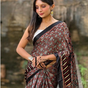 Pu&ograve; includere: Un sari nero caratterizzato da un motivo geometrico ripetitivo di quadrati rossi, bianchi e verde acqua con un bordo nero. Il tessuto presenta un design stampato con il logo KUTCH IQ VARS A BANJARA COUTURE visibile sulla parte drappeggiata.